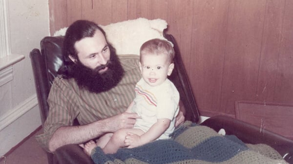 A father with a large beard holding and looking at his son, who is sitting on his lap.