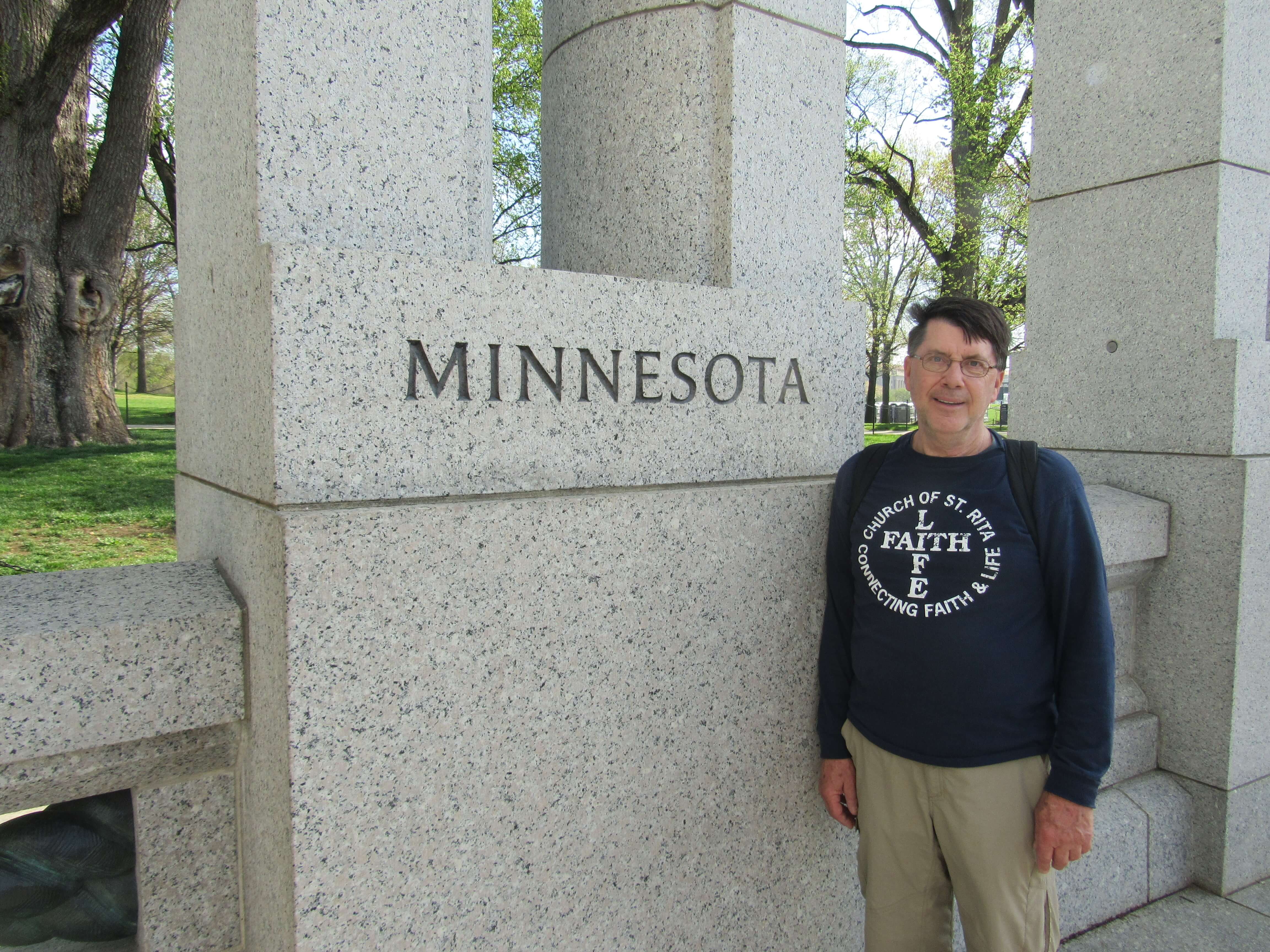 Contemporary photo of an older man at a granite memorial or building that has "Minnesota" carved into it at head height.