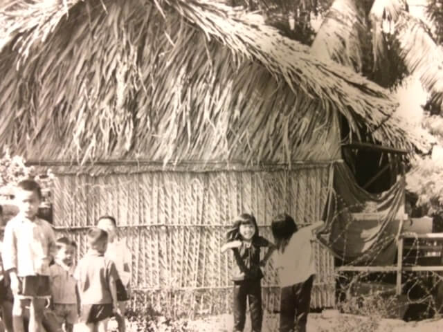 Black and white photo of a group of Vietnamese children playing in front of a thatched hut.
