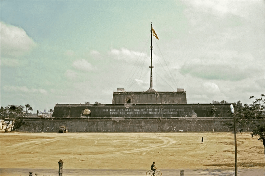 A large, drab building rising out of a desolate landscape. A South Vietnam flag waves high above the building.