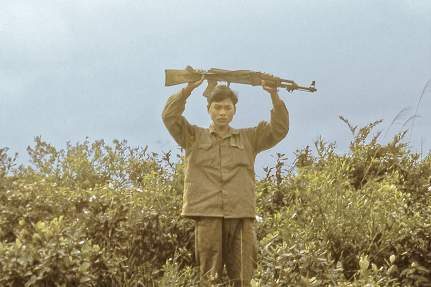 A Vietnamese soldier standing among bushes with his gun raised above his head, potentially surrendering.