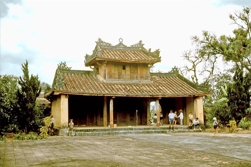 Exterior of a simple two-storied Asian temple, surrounded by greenery. Asian people sit and stand on the steps.