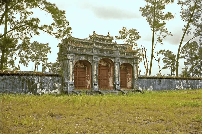 Exterior of a simple Asian temple, surrounded by overgrown greenery.