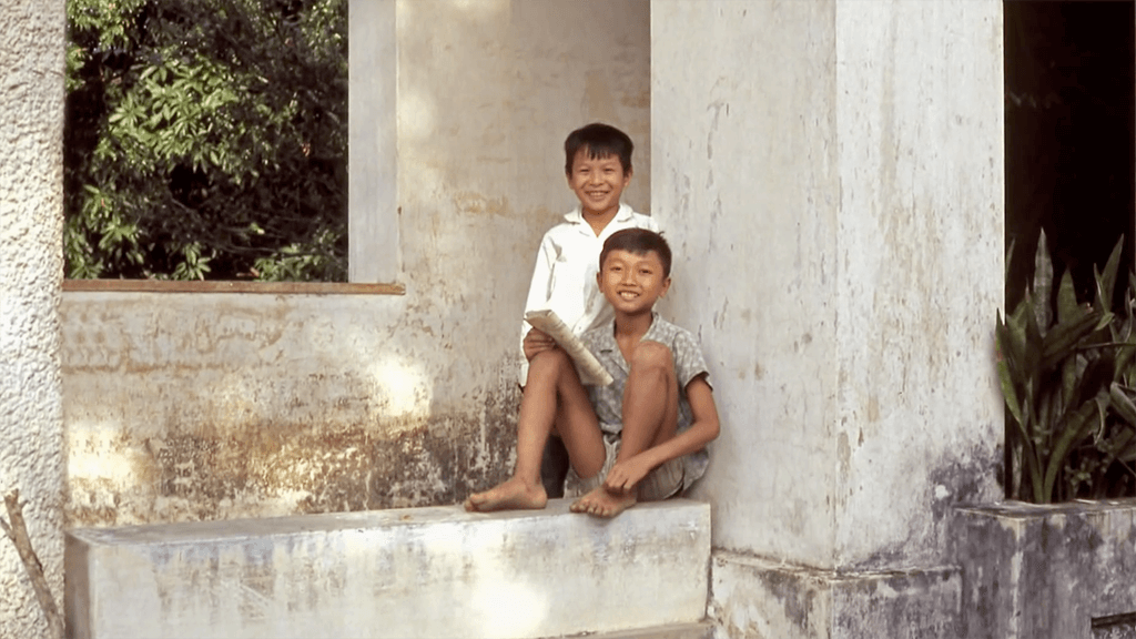 Two smiling Asian boys, sitting and standing outside a white building.
