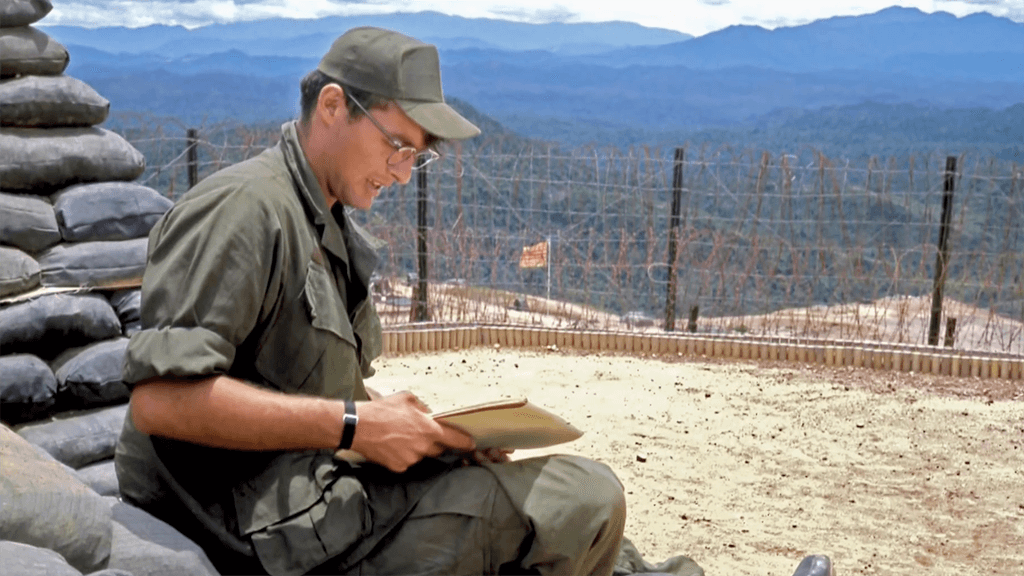 A soldier wearing glasses and a cap, sitting on sandbags on top of a mountain, holding a note pad.