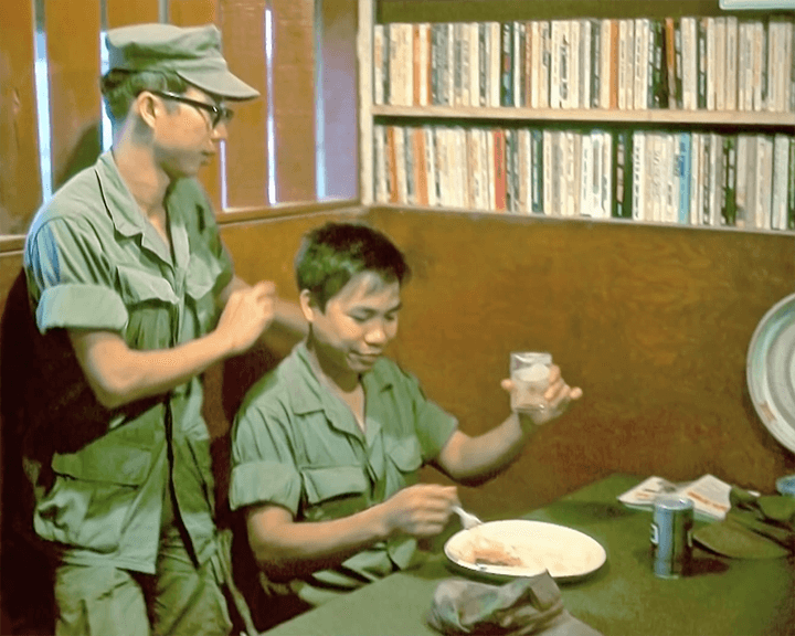 Two Asian men in uniform. One is eating and drinking, the other one is standing behind him.
