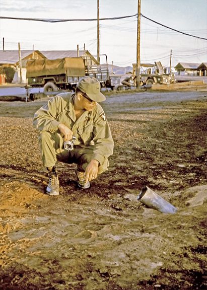A soldier with a camera in hand bent down to look at an exploded rocket in the ground.