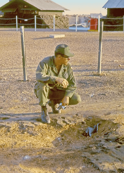A soldier with a camera in hand bent down to look at an a spot in the ground where presumably a rocket or grenade blew up.