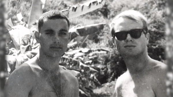 Two young, shirtless men in a jungle. The one one the right is wearing sunglasses.