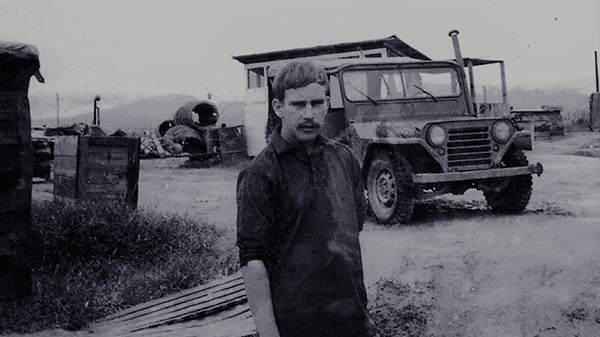 A somber looking young man standing outside, a Jeep in the background.