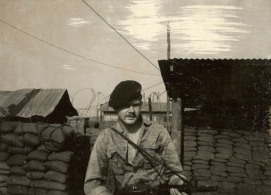 A young US soldier in a beret amd holding a rifle, standing outside an encampment with corrugated tin roofs, razor wire, and sandbags.