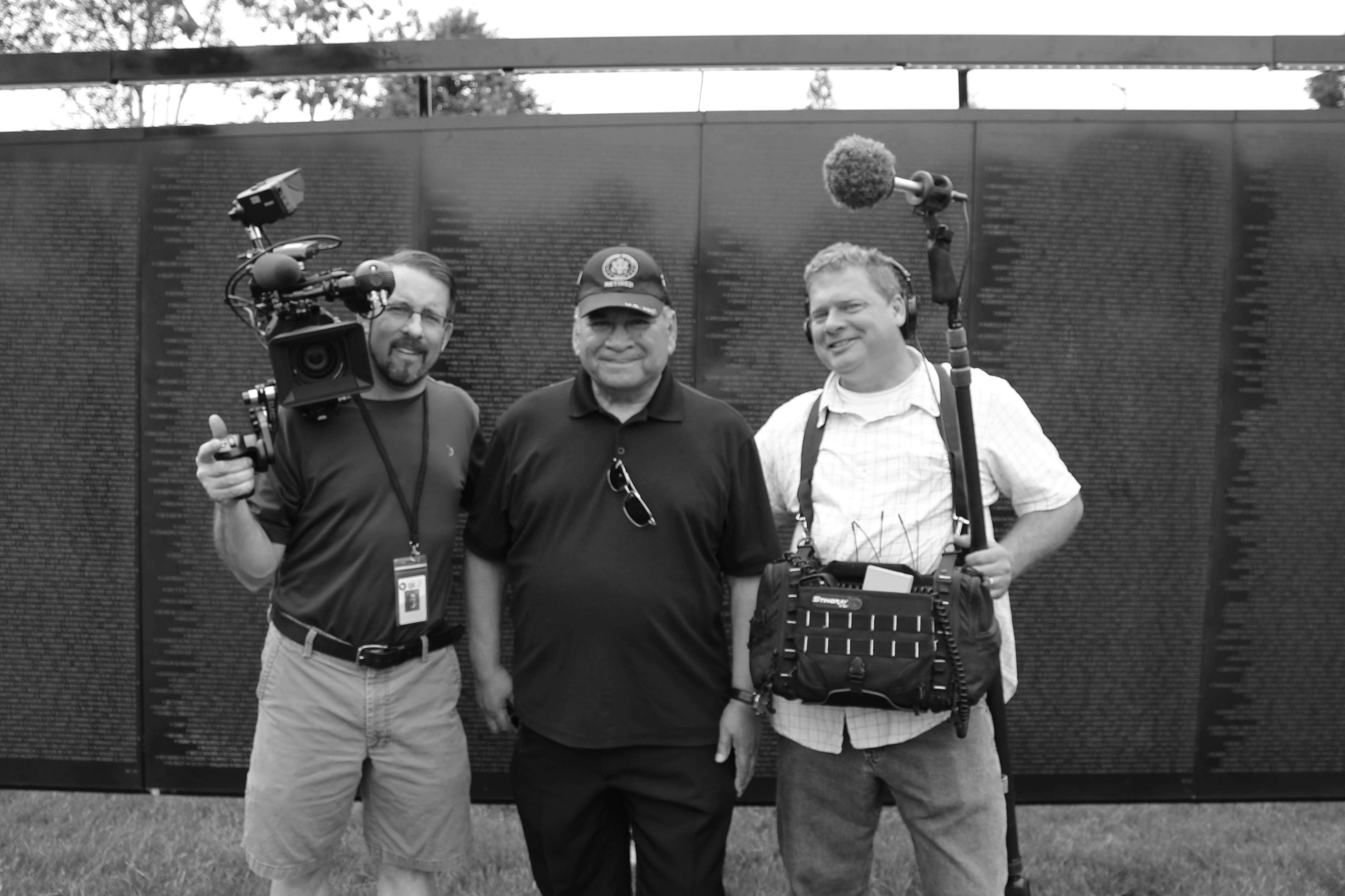 Modern day black and white photo of two middle aged men with camera and audio gear flanking an older gentleman dressed in black with a Vietnam veteran cap on. They are standing in front of a scaled replica of the Vietnam Wall.