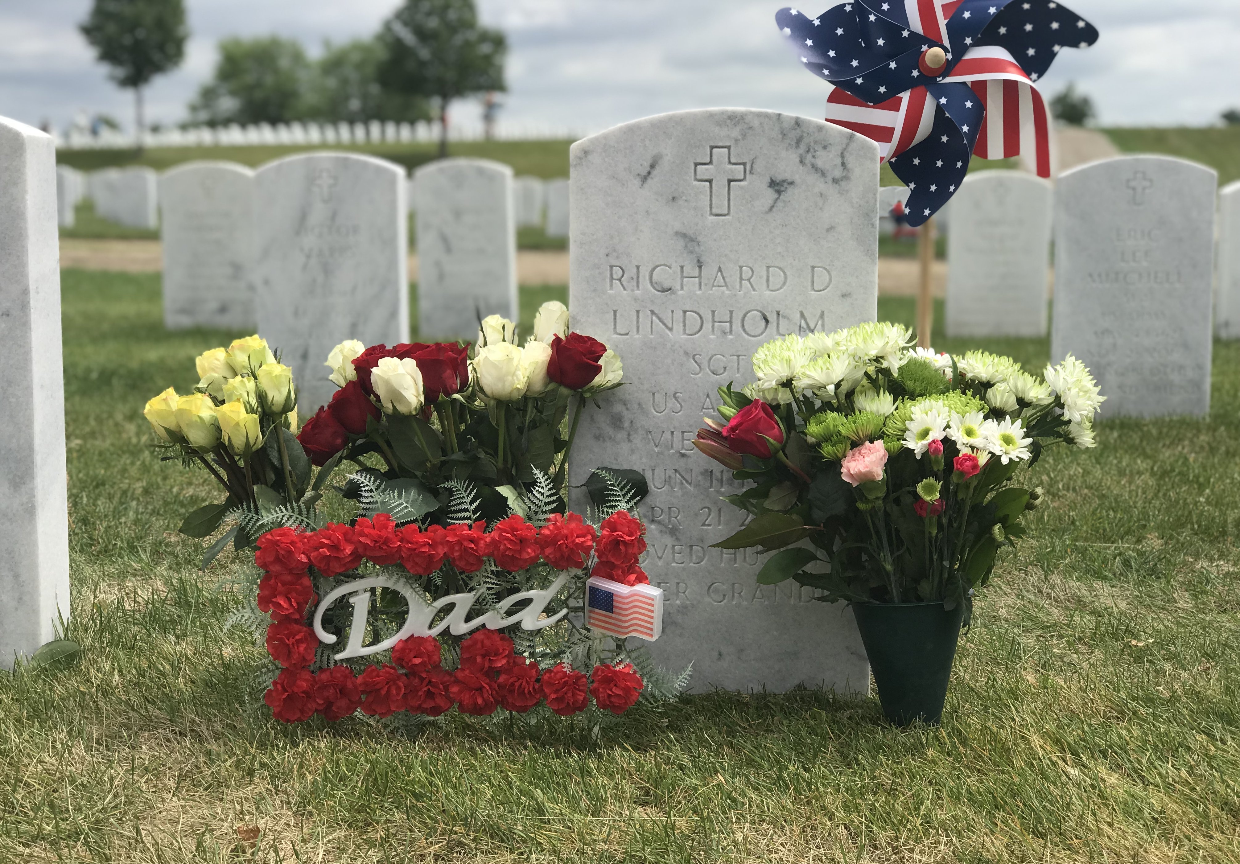 An official military grave marker for Richard D Lindholm, with flowers and an American flag pinwheel.