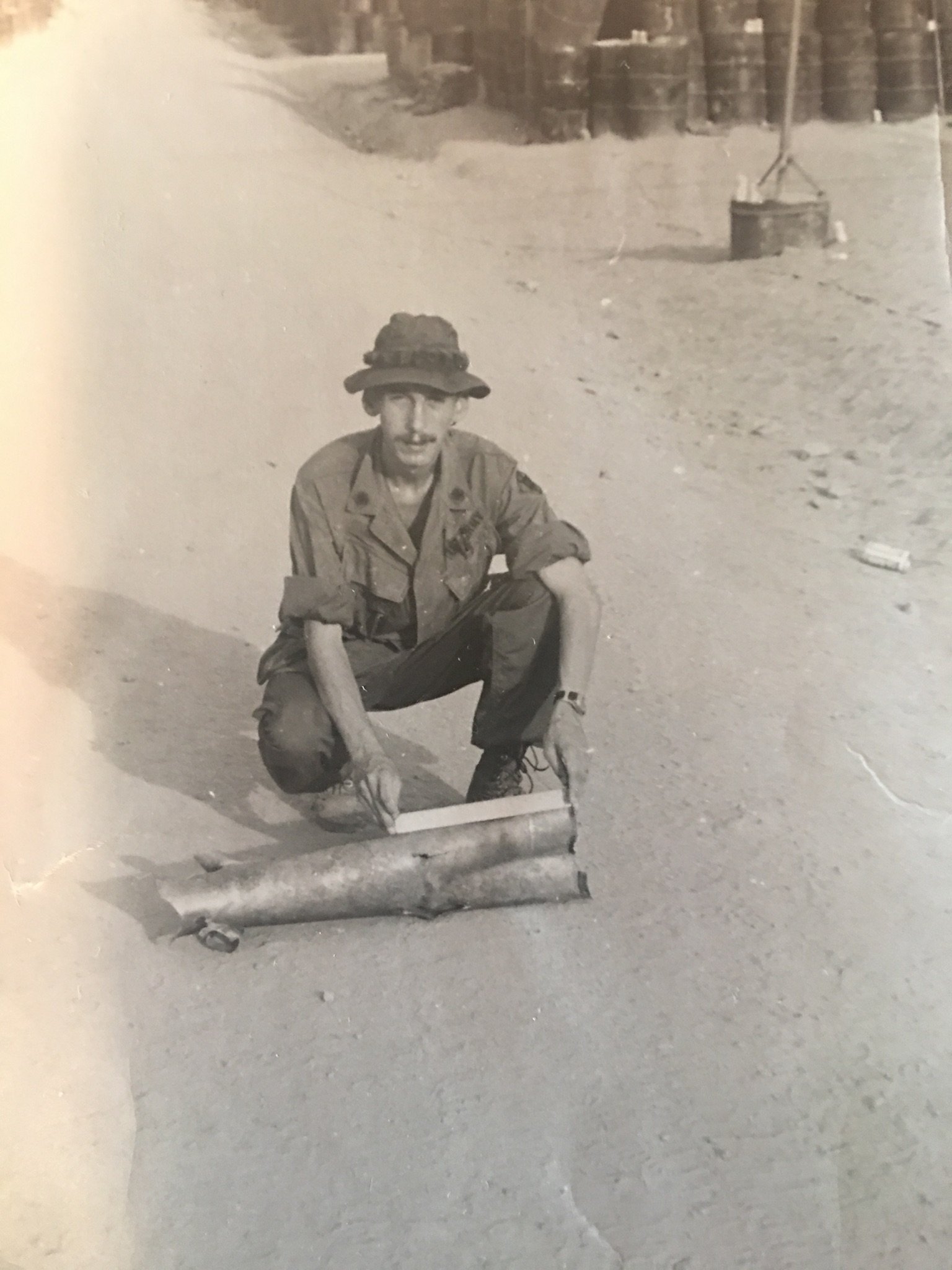 US soldier in a boonie hat knelt down next to an exploded rocket.