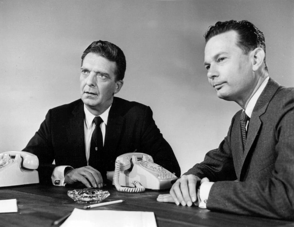 Two middle-aged men sitting at a table, each with an old desk phone, and with an ashtray sitting between them.