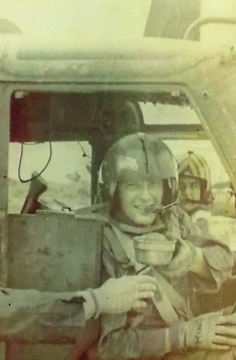 A US soldier in a helmet, sitting in a cockpit, handing off or taking a cup of something to or from someone who is out of frame.