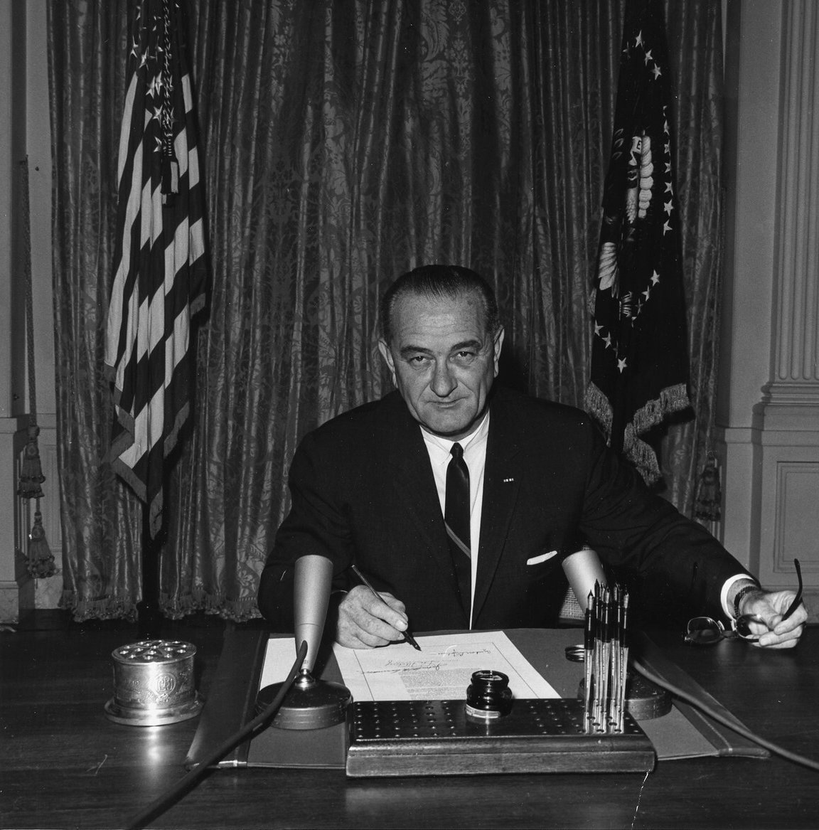 LBJ sitting at a desk and looking at the camera, right hand holds a pen and left hand holds his reading glasses.
