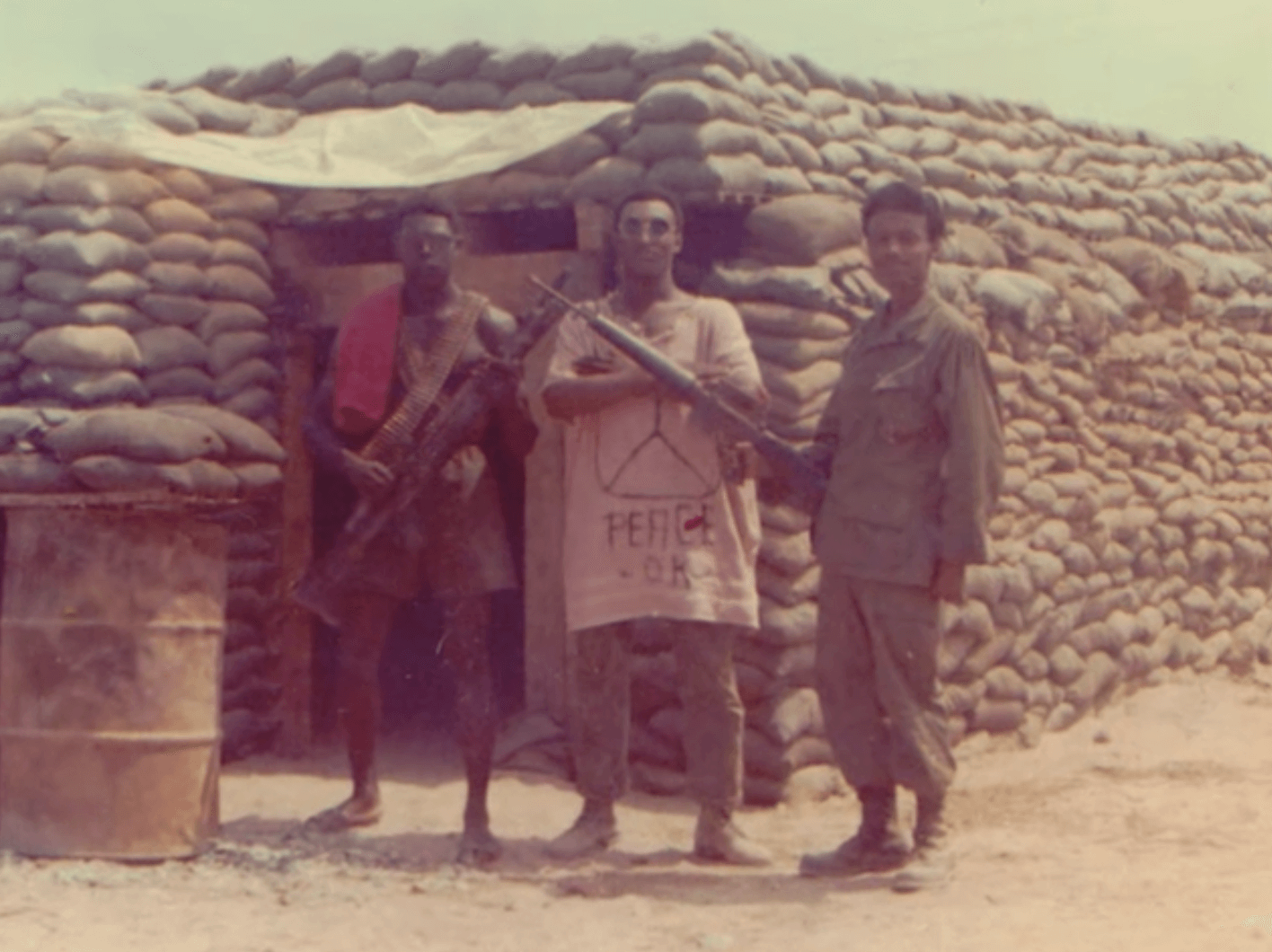 Three black soldiers standing outside a sandbag bunker. The leftmost is in shorts, holding a gun and with bandoliers strapped across his body; the middle wears an oversized t-shirt with a peace sign that says "Peace OK"; the rightmost is in full gear, also holding a gun.