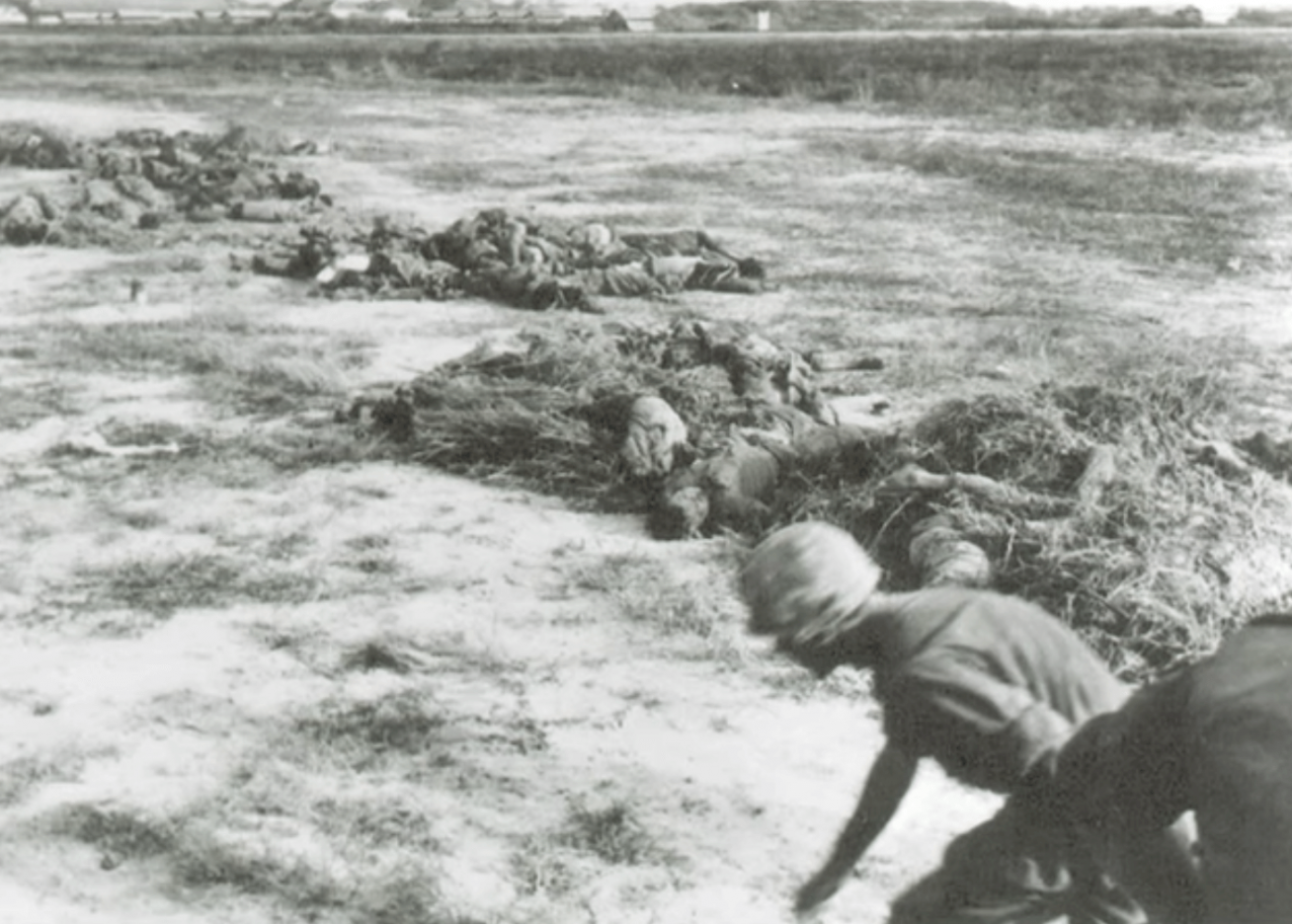 A soldier running and ducking in a field. There appear to be bodies on the ground in the background.