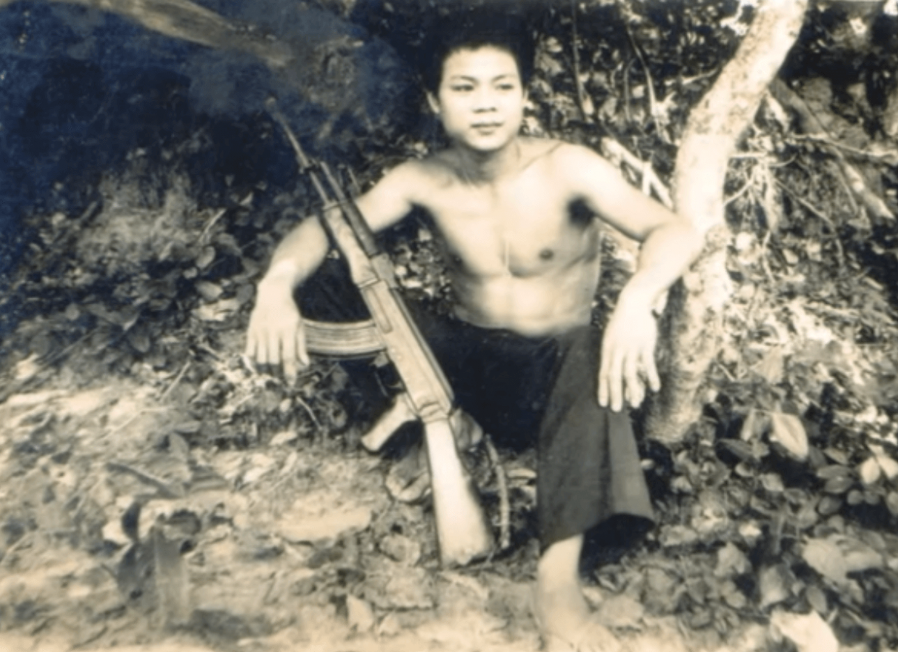 A young Asian soldier, shirtless and in flip flops, sitting down with a rifle propped in his lap.