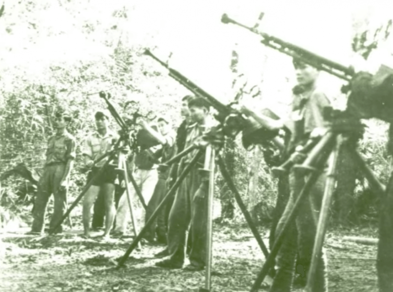 A group of young Asian soldiers standing behind a row of rifles propped up on tripods.