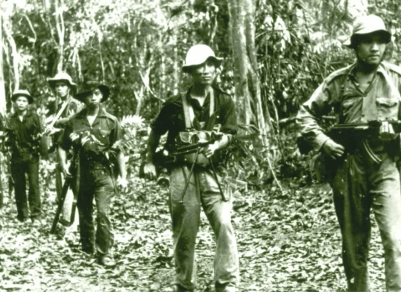 5 Asian soldiers marching in single file through a forested area.