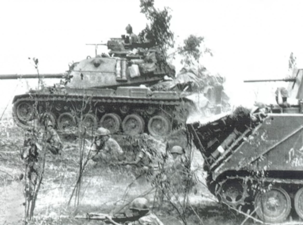 Two tanks moving towards the left frame of the photo, with soldiers on foot in front of and to the sides of the tanks.
