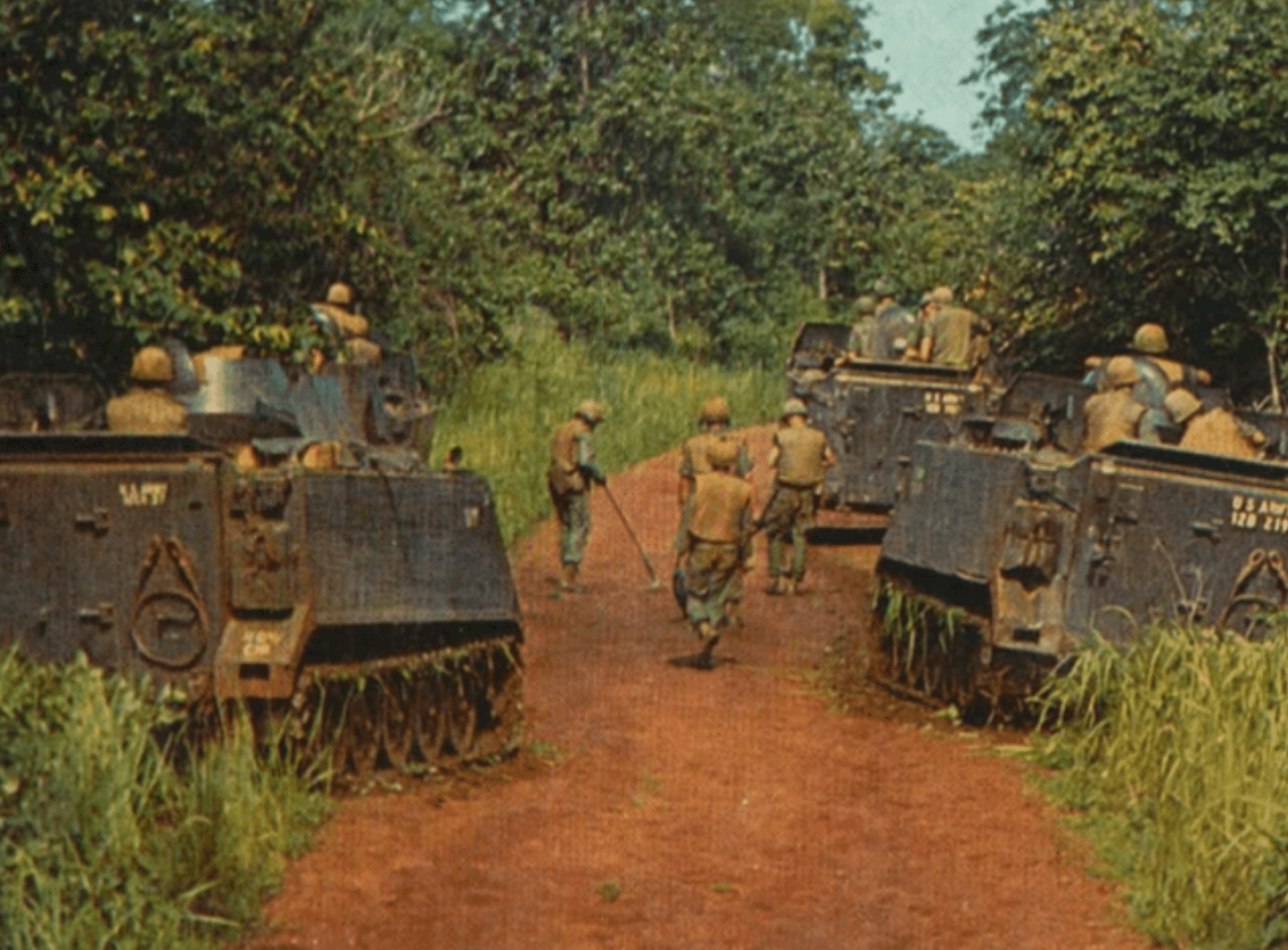 Three tanks on a trail through a wooded area, soldiers patrol on foot between the three.