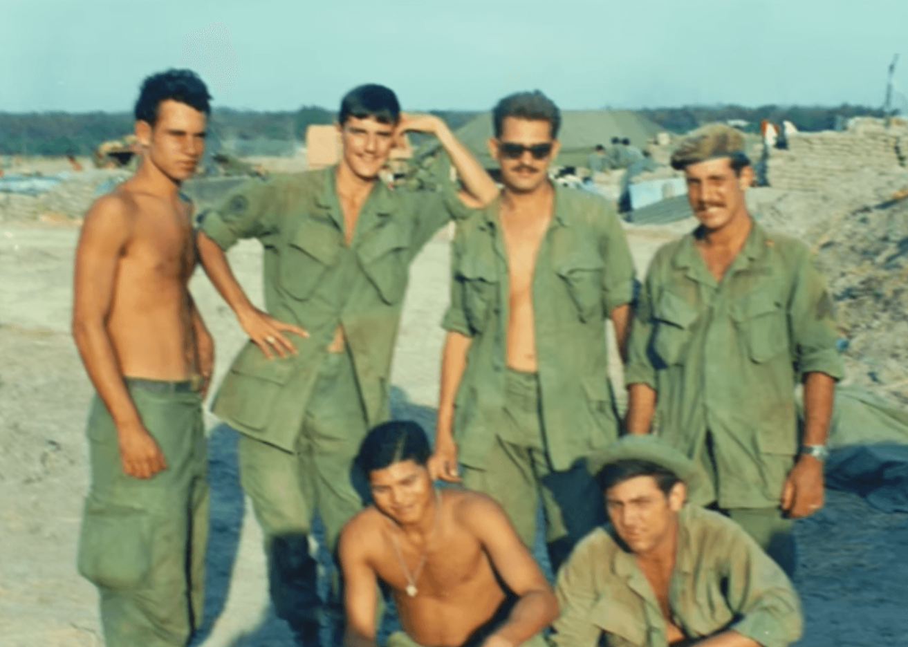 Group of 6 US soldiers posing for a photo. Some are shirtless, some have their shirts unbuttoned.