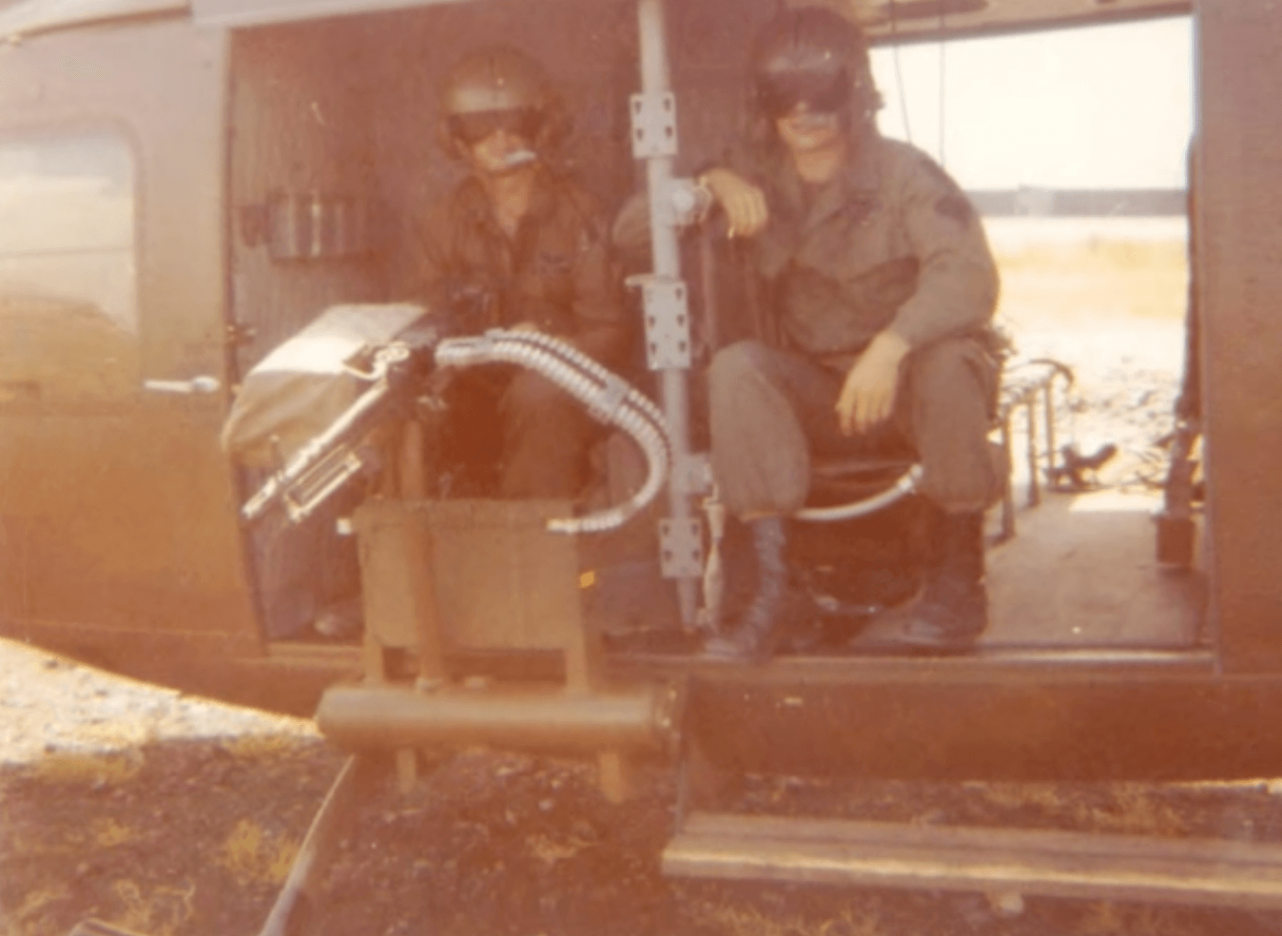 Two pilots with their helmets and shades on, sitting in a Huey helicopter.