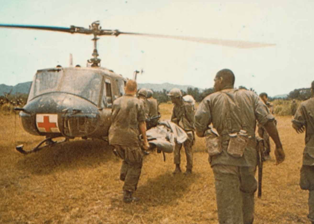 A medevac helicopter with a Red Cross on its nose, soldiers carrying a body on a stretcher toward the helicopter.