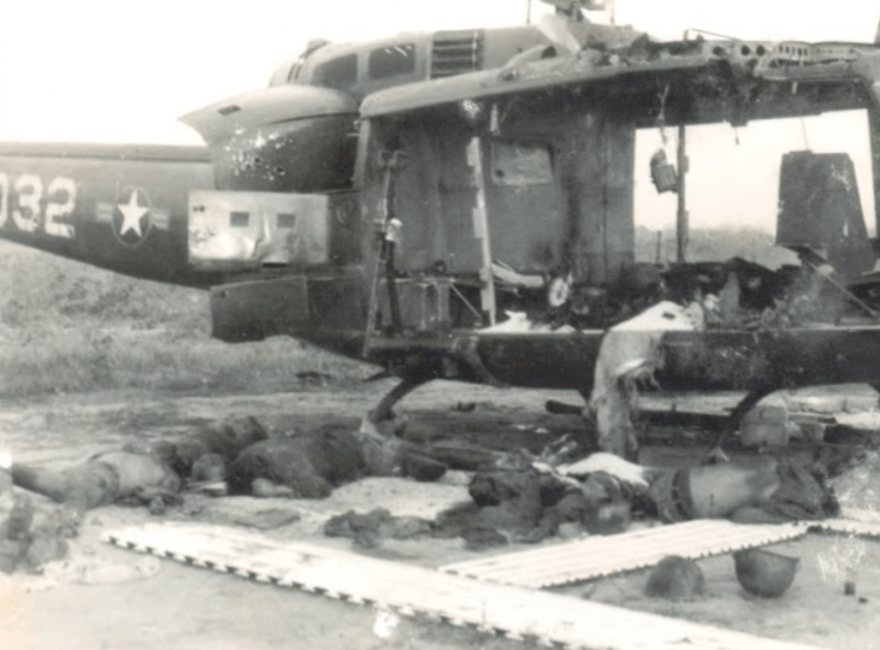 A heavily mortared and bombed out helicopter. There appear to be bodies laying around the ground around the helicopter.