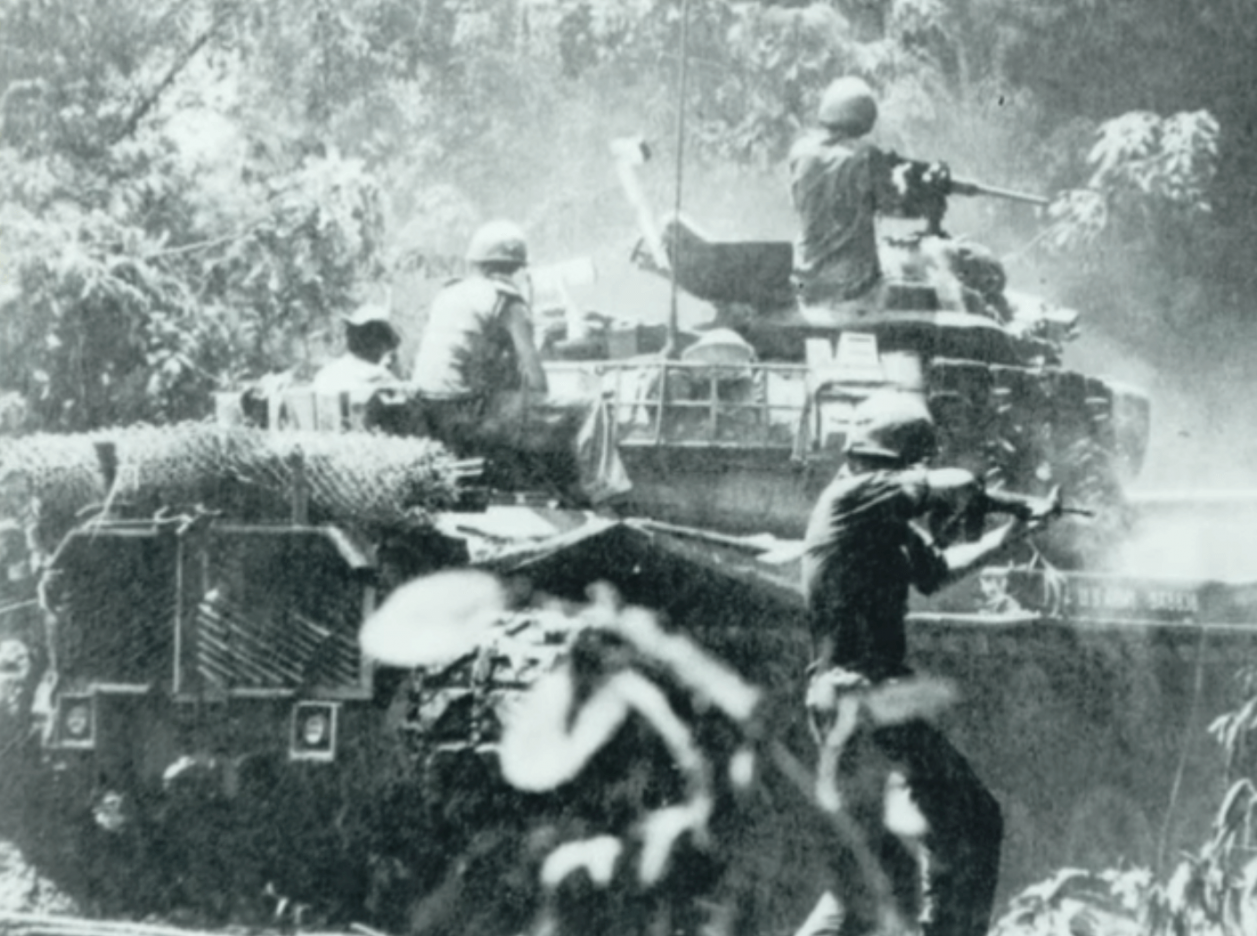 A tank in the jungle with soldiers on top; one soldier is on foot alongside the tank, rifle drawn.