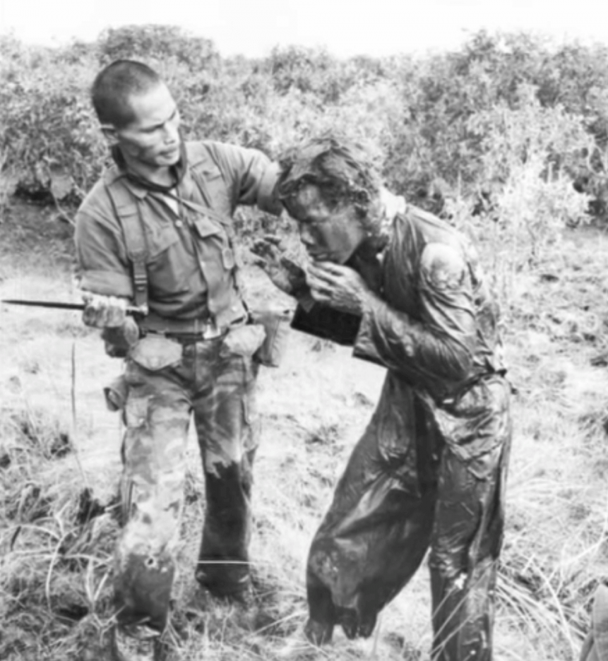 An Asian soldier holding a knife in one hand, and a mud-covered Asian man by the neck in the other.