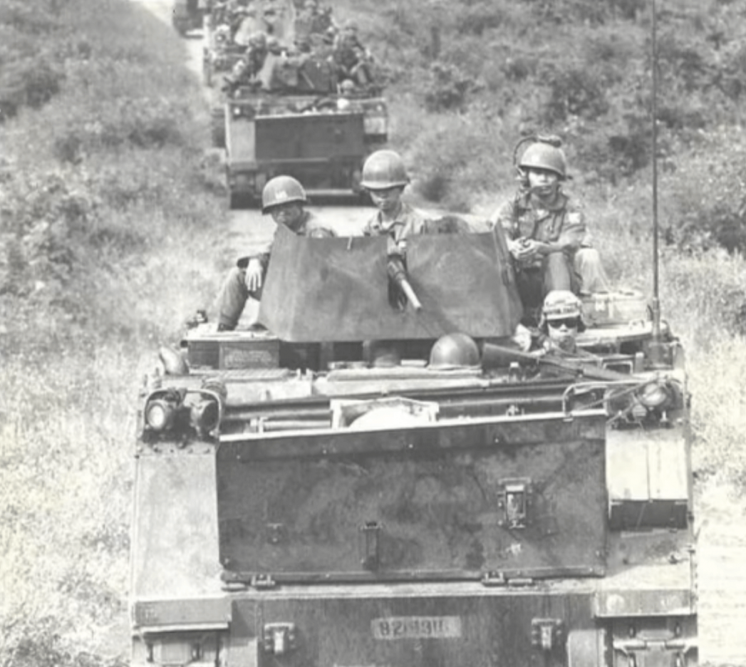 A line of tanks with soldiers atop, approaching toward the camera down a dirt road.