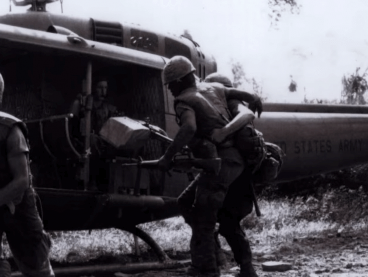 Soldiers loading up a helicopter; one appears to be injured and braced by another.
