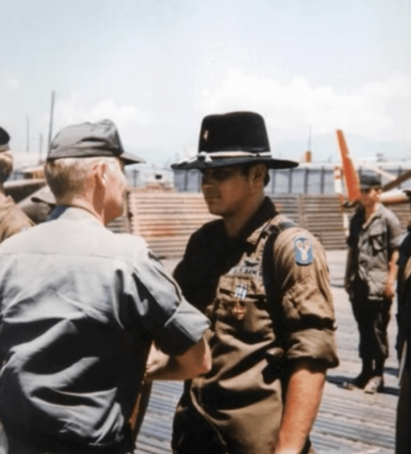 An older soldier shaking the hand of a younger soldier in a cowboy hat, standing in formation among other soldiers.