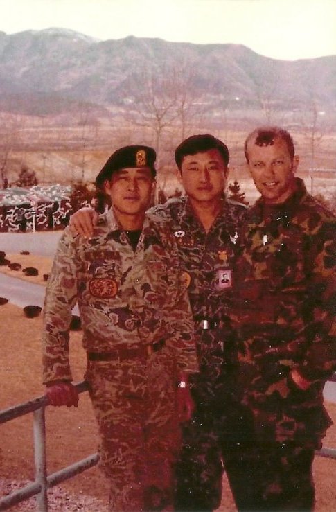 A US soldier posing with two Asian soldiers against some sort of railing; mountains are in the background.