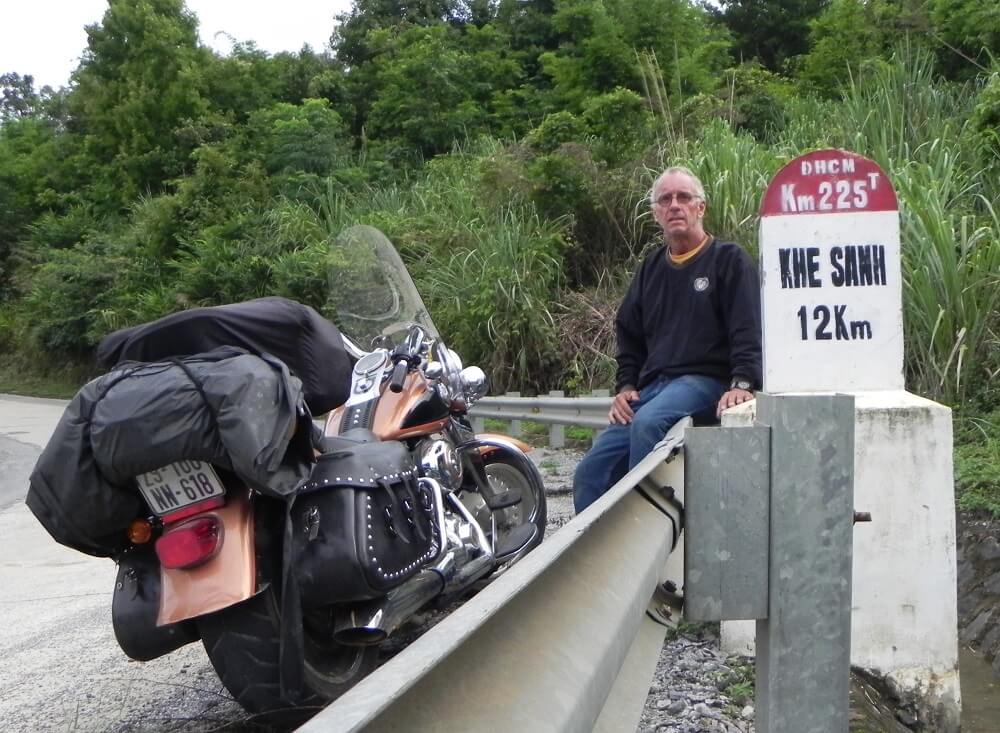 Modern day photo of an older gentleman sitting on a traffic barrier near a sign that says "Khe Sanh 12Km," next to his motorcycle.