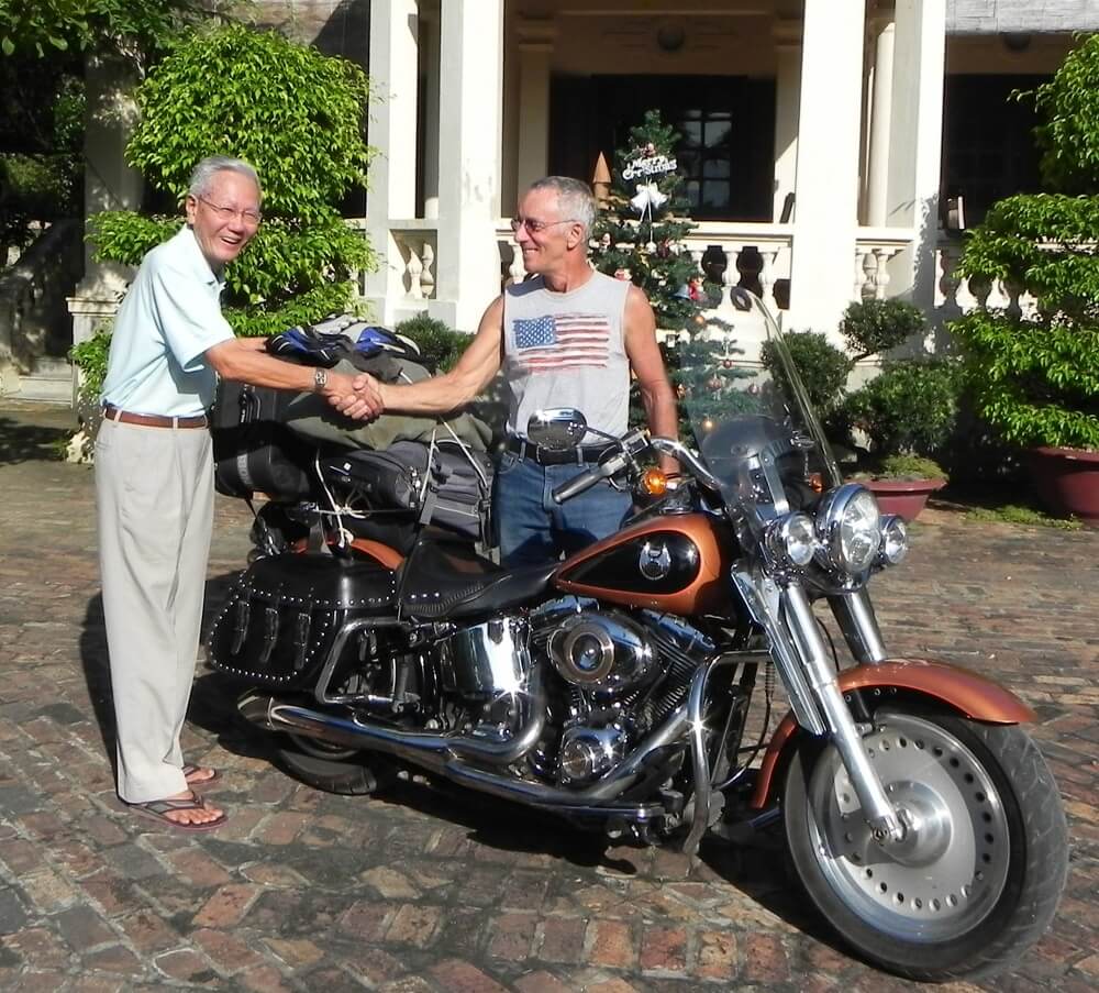 Contemporary photo of an older white gentleman in an American flag sleeveless t-shirt, shaking hands with an older Asian man in slacks and a button down. There is a Harley Davidson between them.