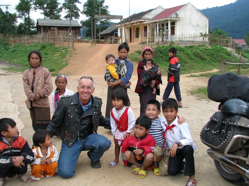 Contemporary photo of an older white man in a leather jacket, crouched down among Asian women and children, on a dirt road.