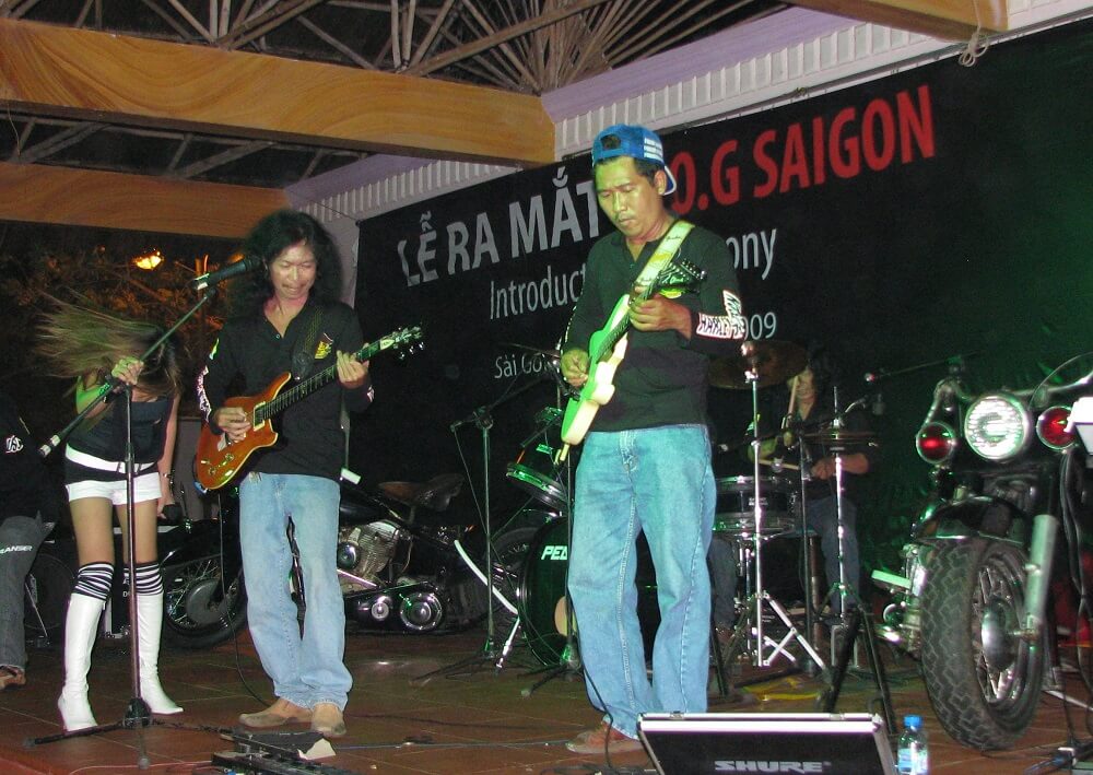 Contemporary photo of an Asian rock band playing on an indoor stage. There are motorcycles in the background.