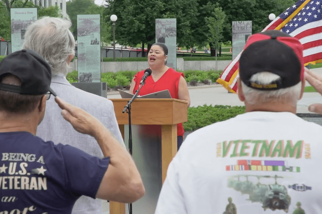Woman in a red shirt singing at a podium while veterans salute.