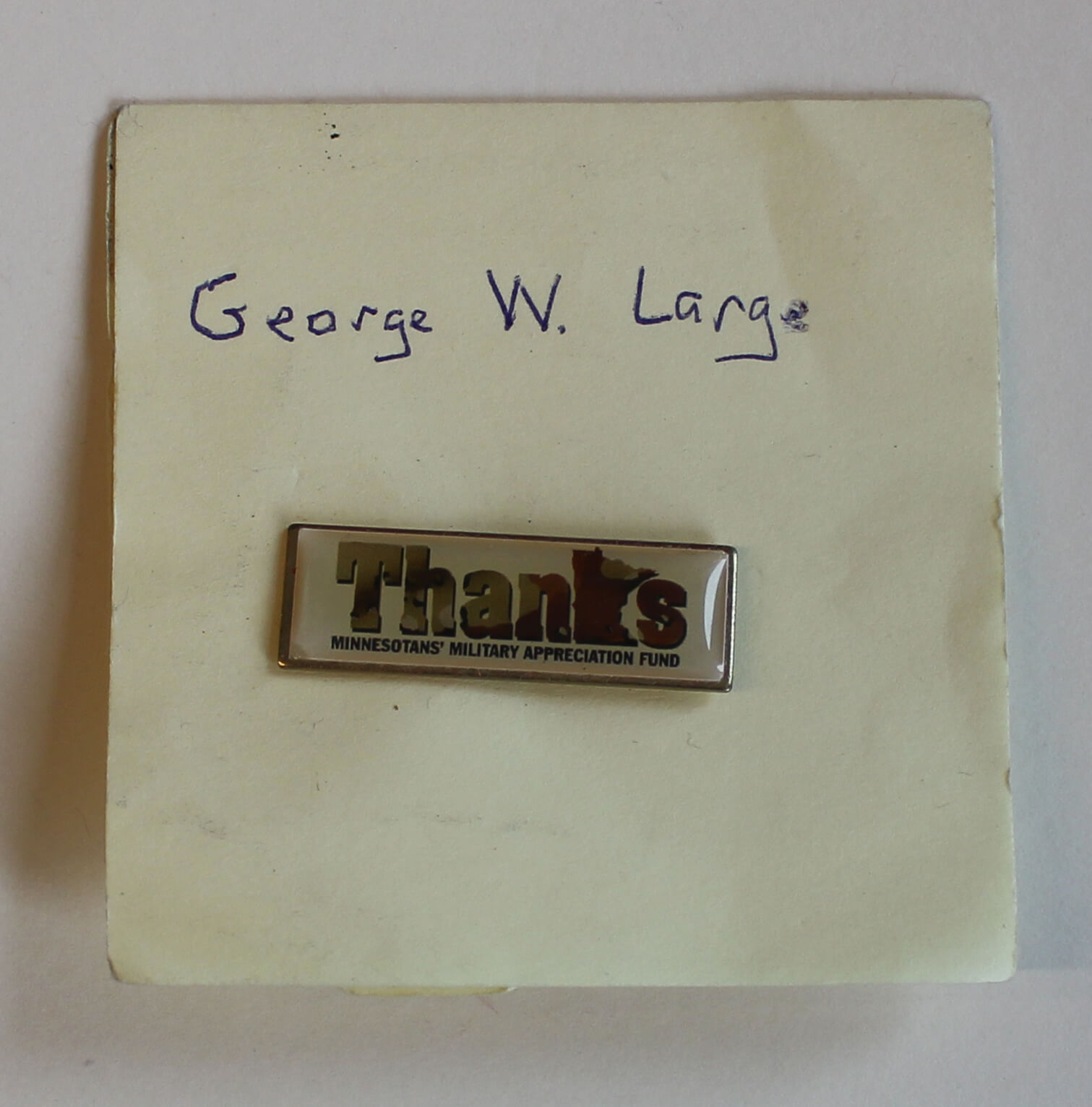 Yellow Post-It note with the name George W. Large written on it with a Thanks- Minnesotan's Military Appreciation Fund pin.
