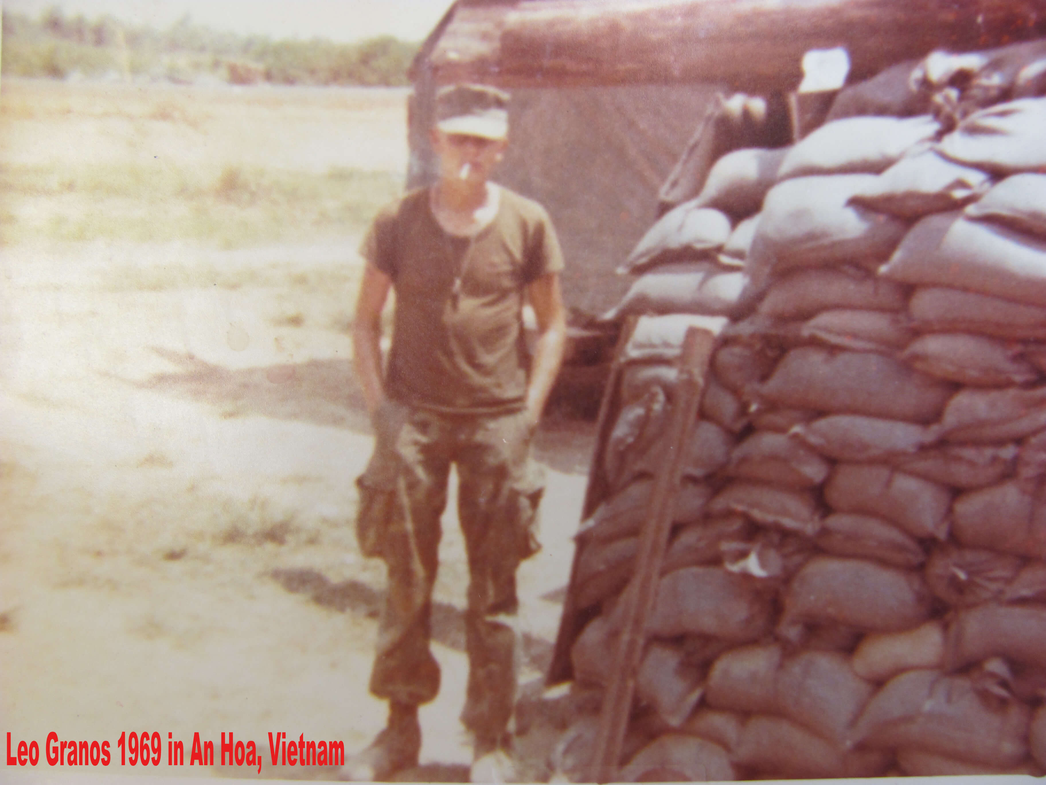 A young soldier standing near a mound of sandbags with a cigarette in his mouth and his hands in his pockets. Text on photo reads "Leo Granos 1969 in An Hoa, Vietnam."