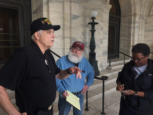 Man in a Vietnam veteran hat talks to a man and a woman in front of a stone building facade.