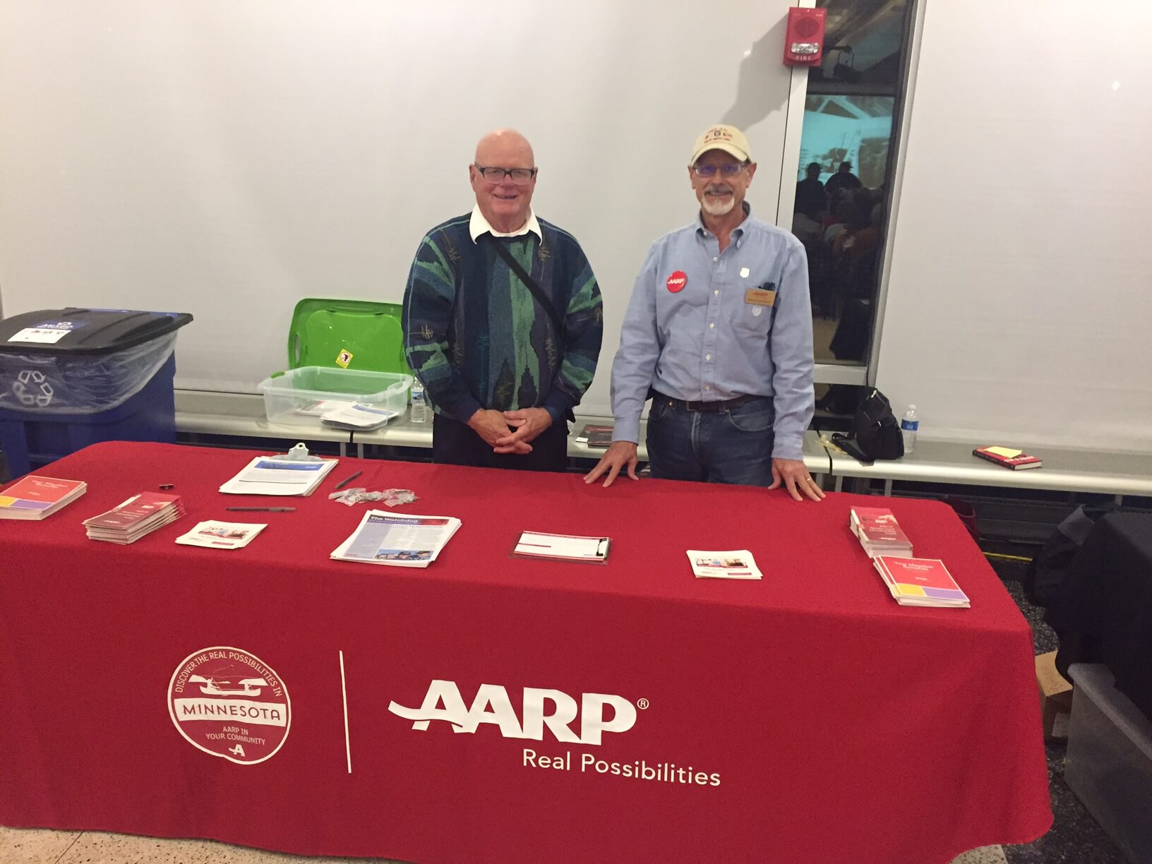 Two men standing behind a red AARP table at an event