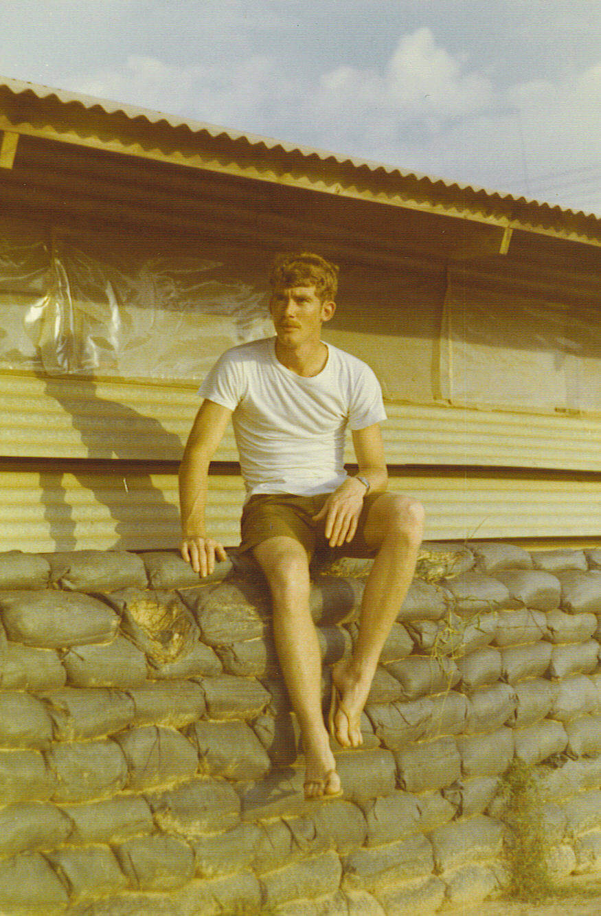 Soldier in a t-shirt and shorts sitting on top of sandbags in a military camp